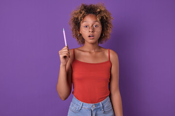 A thoughtful young woman in a red top holds a pencil, gazing inquisitively with a vibrant purple background enhancing her expression.