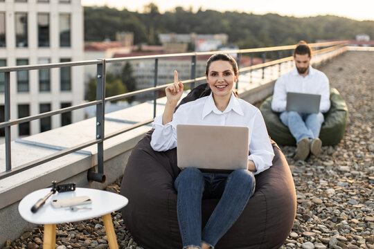 Two people working remotely on rooftop using laptops and sitting on bean bags. Outdoors office setup promoting remote work and modern workspace concepts, with focus on productivity and flexibility.