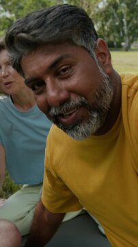 Vertical UGC handheld shot of Biracial middle-aged man filming video for vlog with Caucasian senior friends or having video call, talking after outdoor practice in park on summer day
