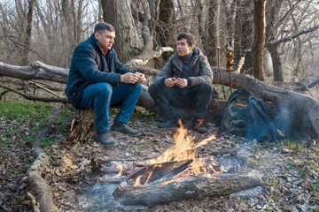 Two men make a campfire for cooking in the forest, father and son sitting on a log while hiking and outdoor activities, early spring landscape