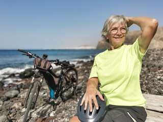 Fototapeta premium Happy senior sporty woman stops after a bike ride on the beach and takes off her helmet. Sunny day and horizon over the sea