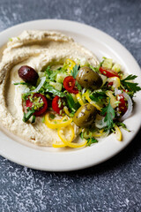 traditional hummus loaded with  fresh cucumbers, olives, tomatoes, pepper, onions and herbs on plate on dark grey table, flat lay, close-up