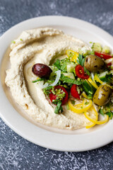 traditional hummus loaded with  fresh cucumbers, olives, tomatoes, pepper, onions and herbs on plate on dark grey table, flat lay, close-up