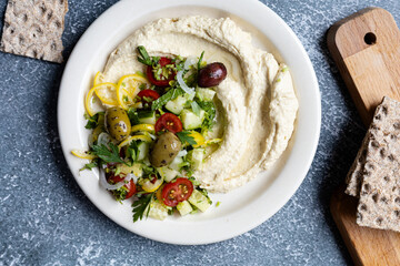 Traditional hummus served with olives, cucumbers, tomatoes, pepper and herbs on dark bachground, close up
