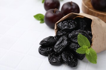 Paper bag with delicious prunes, mint in and fresh ripe plums on white tiled table, closeup. Space...