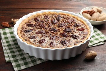 Delicious pecan pie in baking dish and fresh nuts on wooden table, closeup