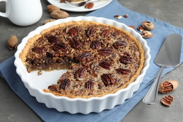 Delicious pecan pie in baking dish, cake server and fresh nuts on gray textured table, closeup