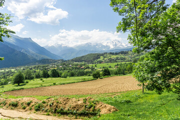 Obraz premium Plowed field, green grass, bushes and trees, village. Snow-capped mountains and peaks in the background