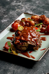 A juicy steak with a rich, red sauce, served with roasted potatoes and cherry tomatoes on a gray plate.
