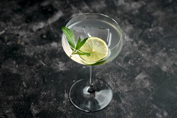 A classic mojito cocktail in a coupe glass with mint leaves and a lime wedge. The drink is served on a dark, moody background.