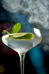 A classic mojito cocktail in a coupe glass with mint leaves and a lime wedge. The drink is served on a dark, moody background.