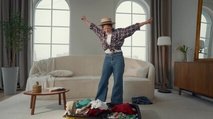 Happy woman traveler packing suitcase and dancing in room, waiting for summer. Cheerful girl in straw hat has fun and joy when unpacking travel bag in hotel room, enjoying travel and weekend trip