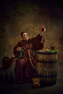Man, medieval monk lifting glass of beer, pint of ale against dark vintage background. Rich legacy of abbey brewing traditions. Concept of comparison of eras, history, brewery, traditions, festival