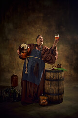 Medieval monk, man in period clothes with beer barrel and pint raising glass of ale against vintage...