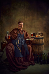 Naklejka premium Medieval monk, holding pint of ale and plate of food, with wooden barrel filled with various drinks of beer against vintage background. Abbey ale. Concept of comparison of eras, history, brewery