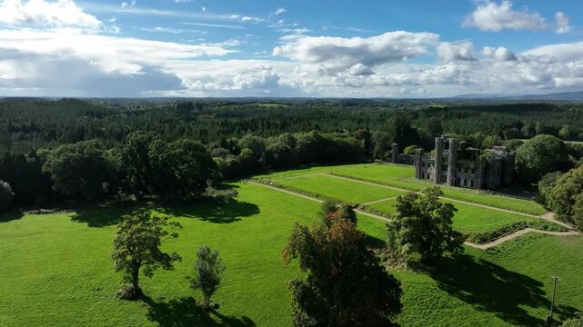Castle Saunderson, County Cavan, Ireland, September 2022. Drone orbits clockwise, wide aerial view of old grey castle with garden in front, surrounded by lush green trees and cloudy sky overhead.