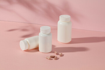 A few white medicine jars without label displayed on top of pink table with some vitamin capsules flat lay beside. The photo was taken from high angle, blank space for designing