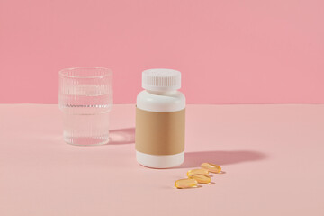 The unique promotional template photo from frontal shot over pink background. A white jar with blank brown label placed next to some yellow capsule and a glass of water, copy space