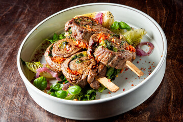 A plate of rolled and stuffed meat slices garnished with herbs, on a bed of lettuce, accompanied by onion rings and spices. Perfect for culinary presentations and food photography.