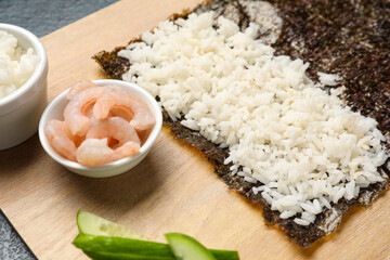 Wooden board with nori and ingredients for preparing sushi rolls on dark background, closeup