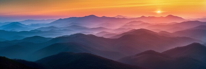 Fototapeta premium The mountains are covered in a misty blue haze. The sun is setting in the distance, casting a warm glow over the landscape. The sky is a mix of orange and pink hues, creating a serene
