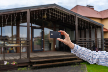 Hand holding plastic credit card with new house at background. Broker, mortgage, property, real estate concept. Buying or renting