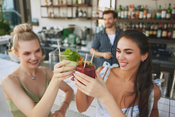 Cheers. Two smiling beautiful women clinking glasses with cocktails in modern bar settings, with bartender on background. Happiness and joy. Friendship, party, leisure, celebration, relaxation concept