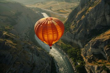 Obraz premium Bright orange hot air balloon floats peacefully above a winding river cutting through a rugged canyon, offering a breathtaking view of the natural beauty below