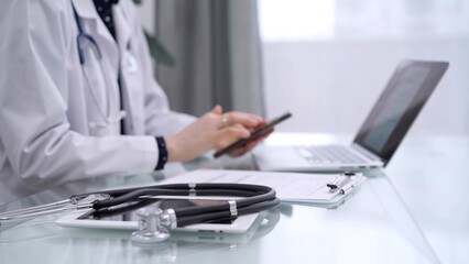 Stethoscope and clipboard are lying on the glass table while doctor woman is using a smartphone at the background. Medicine concept
