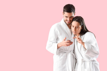 Happy young couple in robes with mobile phone on pink background