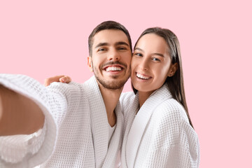 Happy young couple in robes taking selfie on pink background