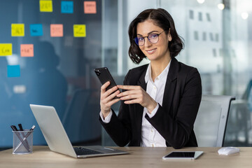 Confident businesswoman in glasses using phone in modern office. Smiling professional working on laptop surrounded by colorful sticky notes, showcasing productivity and modern business environment.