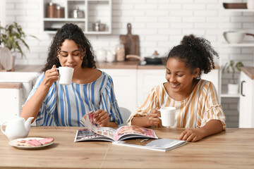 Beautiful young happy African-American mother with her daughter drinking tea and reading magazine in kitchen at home