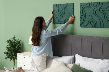 Young woman hanging picture on green wall in bedroom