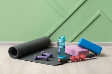 Sports bottle of water with different equipment and yoga mat on floor near green wall
