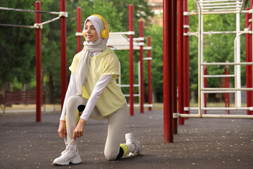 Sporty Muslim woman in headphones tying shoelaces outdoors