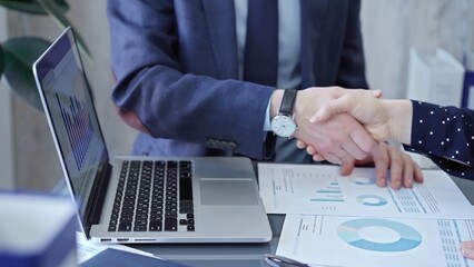 Two business people professionals shaking hands in office setting, sealing a deal with laptop and financial graphs visible. Business handshake over laptop and reports