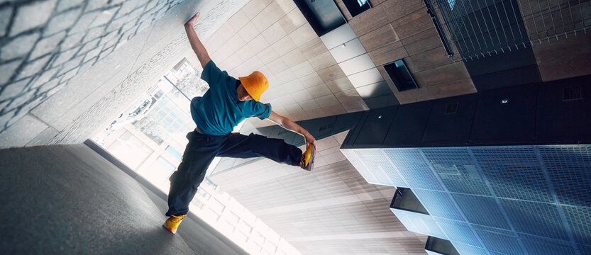 Creative photo. Dancer in yellow bucket hat and blue shirt performs handstand against modern urban backdrop. Strength and style. Concept of lifestyle, extreme sport, freestyle, activity, motion. Ad
