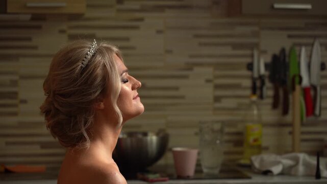  A bride with closed eyes and a tiara on her head puffs up her cheeks playfully while sitting in a kitchen, captured in a candid moment.