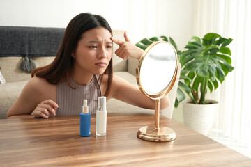 A woman looks at her reflection in the mirror with a concerned expression, possibly worried about her skin. The bright room and natural light emphasize the self-care theme.