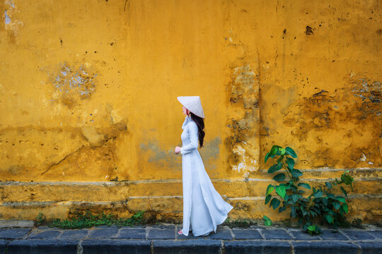 Asian woman wearing traditional Vietnamese culture walks beside an old wall in Hoi An, Vietnam.