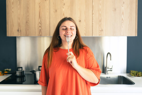Portrait of excited hungry young woman holding fork in her mouth, biting and dreaming about delicious healthy food at the kitchen. Diet concept