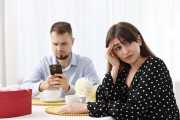 Embarrassing date. Bored woman sitting at table with man using smartphone indoors, selective focus