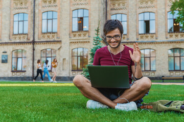 Young male student man freelancer guy in earphones sitting near study university campus using laptop working remote studding online outdoors showing hello gesture has zoom call conversation conference