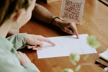 Close-up image of two people reading restaurant menu with QR code. Arms and hands visible, one...