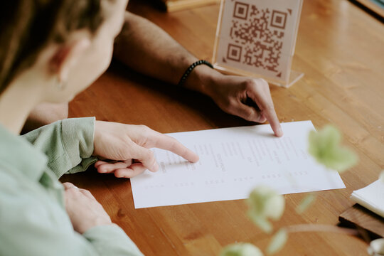 Close-up image of two people reading restaurant menu with QR code. Arms and hands visible, one pointing at menu while other holds it on wooden table