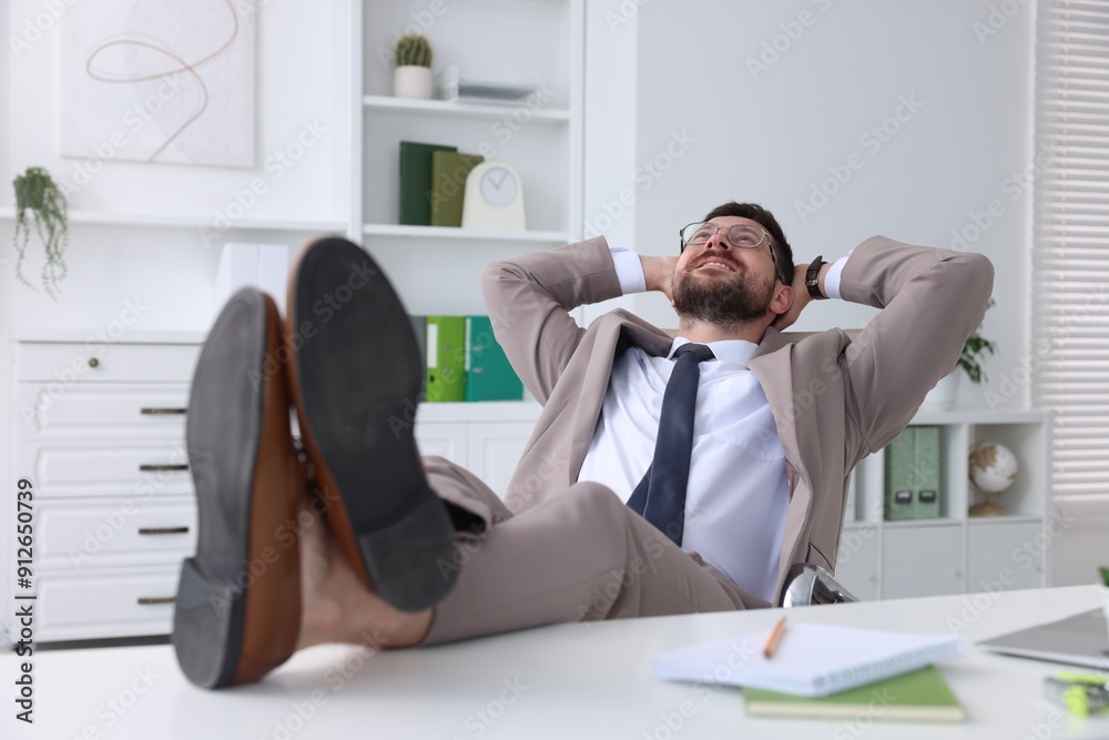 Wall mural Smiling businessman with hands behind his head holding legs on table in office. Break time