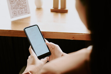 Person holding smartphone while sitting at table, fingers touching screen, with QR code visible in background on wooden surface, creating a serene atmosphere