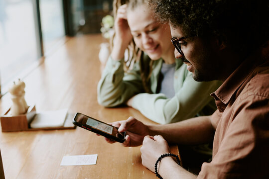 Two people sitting at table in cozy cafe, smiling while looking at smartphone, engaging in conversation. Bright environment with window letting natural light in