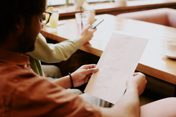 Person in focus holding a cafe menu while another holds a smartphone. Scene depicts relaxed...